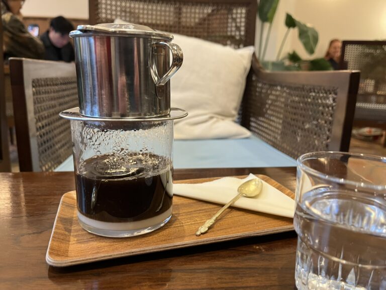 One-person coffee maker on a wooden table, with coffee still dripping through, In the background is a chair with a white cushion on it.