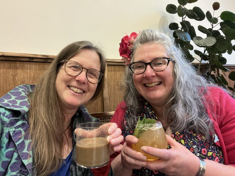 Two women with long hair and glasses smiling into the camera. Both are holding drinks – one looks like coffee and the other a fruity drink.