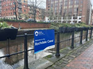 View of a canal and part of the towpath. Blue sign that says 'Rochdale Canal'.