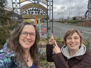 Two women grin into the camera. They are standing in front of a sign saying 'Welcome to Chorley. Lancashire's Market Town'.