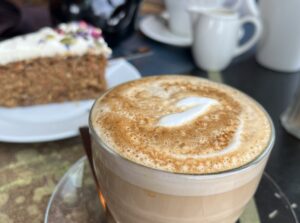 Café table with a frothy cappuccino in a glass cup very close up at the front. Behind are a slice of carrot cake on a white plate, plus a white teapot, cup and milk jug.