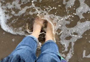 View from above of two legs in blue jeans, with bare feet under water, being lightly splashed by a wave