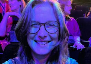 Close up of a woman wearling glasses and bathed in a blue light. She's sitting in an auditorium among other people.