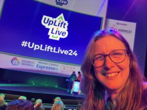 Woman wearing glasses, bathed in orange light. Behind her is a huge screen that says 'UpLift Live' with the hashtag #UpLiftLive24.