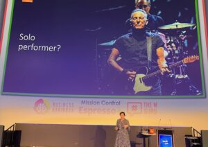 A woman is speaking on stage. Behind her is a huge screen with an image of Bruce Springsteen playing the guitar. The text next to Bruce says 'Solo performer?'