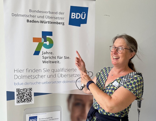 Jane is on the left in a cheesy pose, pointing at the roll-up banner behind her. The banner is proclaiming 75 years of the German Association of Interpreters and Translators (BDÜ).